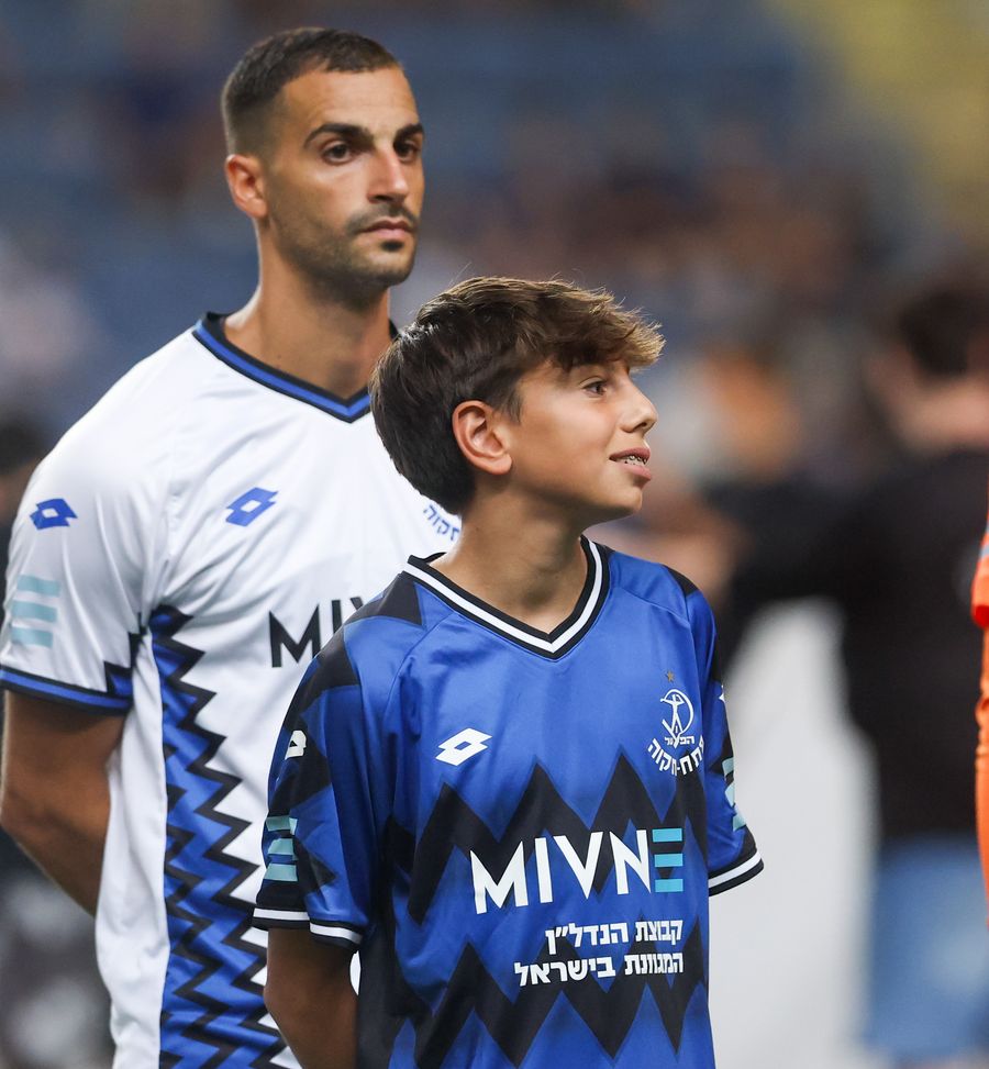 Avishai Cohen (Hapoel Petah Tikva) A young boy in a blue and black patterned jersey stands looking up, with an adult player in a white and blue patterned jersey standing behind him