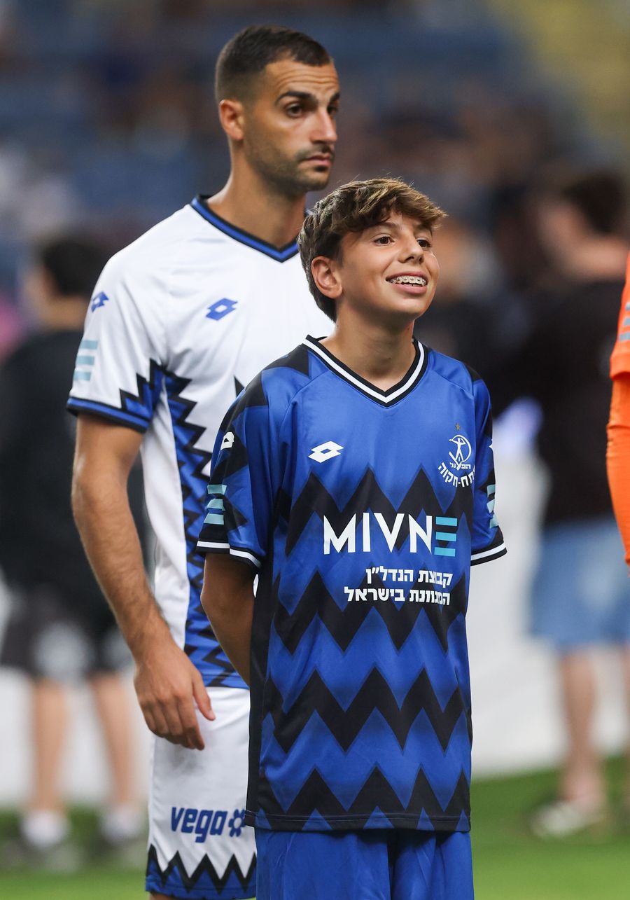Avishai Cohen (Hapoel Petah Tikva) A young boy in a blue and black chevron patterned kit stands smiling, with an adult player in a white kit with blue and black accents standing be