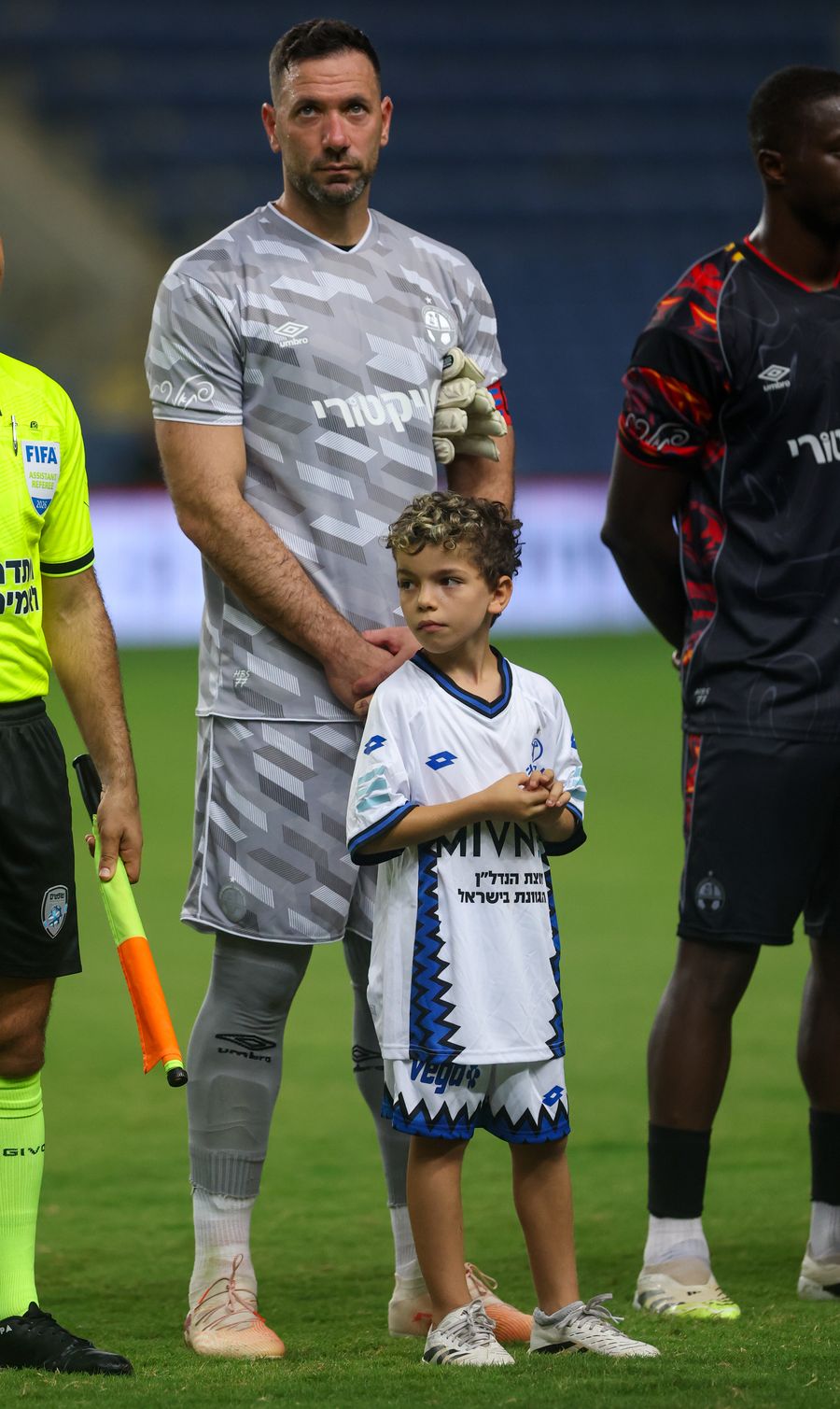 Ofir Marciano (Hapoel Beer Sheva) A young boy in a white kit stands with his hands clasped, next to a goalkeeper in a grey kit.