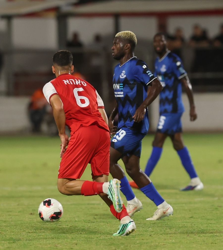 A Bnei Sakhnin player Nino striking the ball with his right foot as a defender attempts to block.