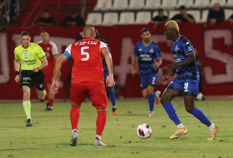 A footballer Player in blue controlling the ball while being challenged by a player in red.