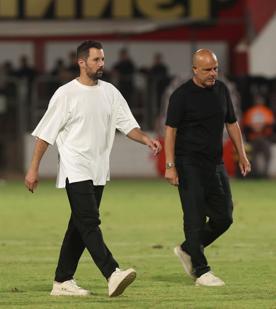 Two men walking across a football field, one in a white shirt and the other in a black shirt