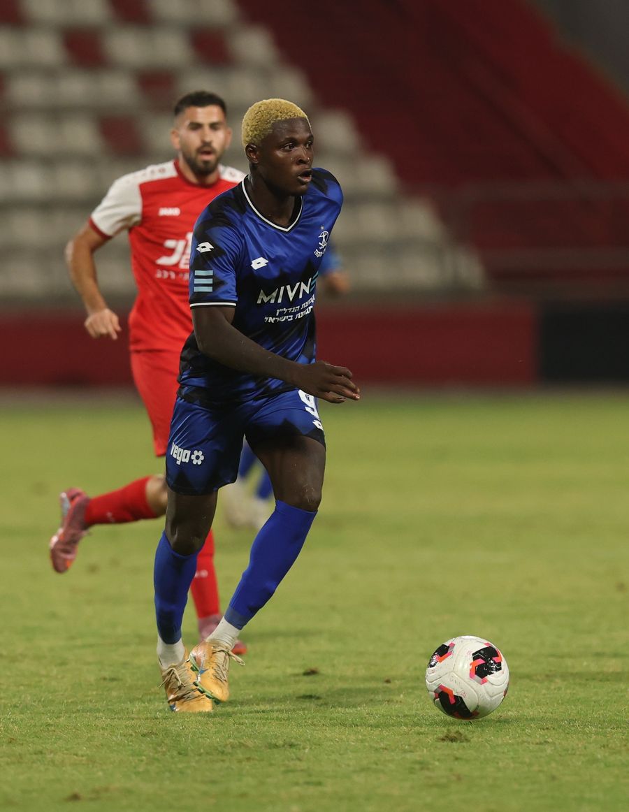 Yves Erick Bile (Hapoel Petah Tikva) runs towards the ball as Basil Khuri (Bnei Sakhnin) runs behind the player in blue