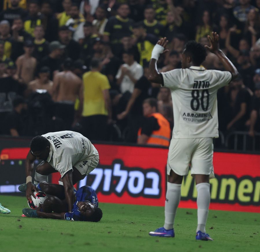 A Hapoel Petah Tikva player is lying on the ground with the ball near him, while another player in white bends over him, and a third player in white raises his hands